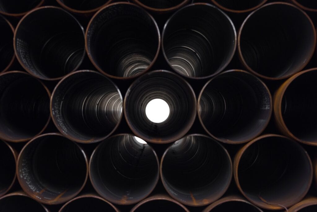 A tightly stacked grid of large, dark metal pipes, with one pipe in the center showing a bright white light at the end of its tunnel.