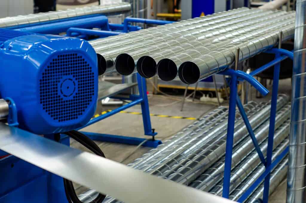 A close-up view of shiny, metallic ventilation ducts, possibly for an HVAC system, being manufactured in a factory. A blue industrial machine is visible on the left.